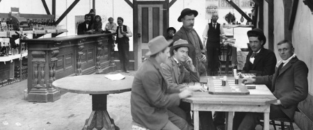 Four men sit around a table playing cards (right foreground). Several people are gathered around one end of a bar (left), including two children sitting on the bar. A barber is set up in the corner with the tools of his trade. Legible signs include: "Pabst Bohemian", "Wagon covers, awnings, made by hand or machine, tents for rent, [...] all kinds [...] of canvas, 136 S. Main St., Los Angeles, Cal.", calendar by "Elcelsior Soda Works [...] Los Angeles, Cal.", "Pabst B Milwaukee", ..."ill[...]e the Barbi"....